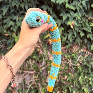 Peut inclure: Un serpent en peluche crochet&eacute; de couleur turquoise douce avec des motifs en forme de losange orange vif le long du corps. Le serpent pr&eacute;sente une t&ecirc;te arrondie, de grands yeux de s&eacute;curit&eacute; en plastique noir et jaune et des joues roses brod&eacute;es.