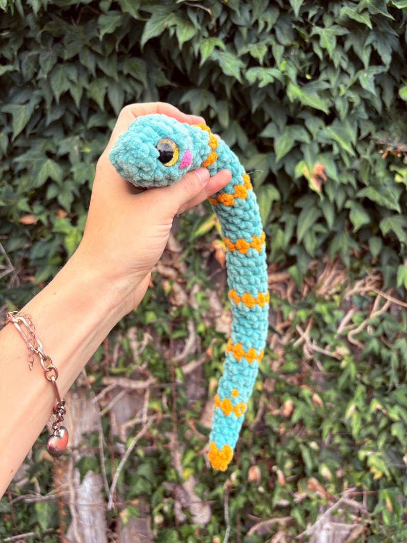 Peut inclure: Un serpent en peluche crochet&eacute; de couleur turquoise douce avec des motifs en forme de losange orange vif le long du corps. Le serpent pr&eacute;sente une t&ecirc;te arrondie, de grands yeux de s&eacute;curit&eacute; en plastique noir et jaune et des joues roses brod&eacute;es.
