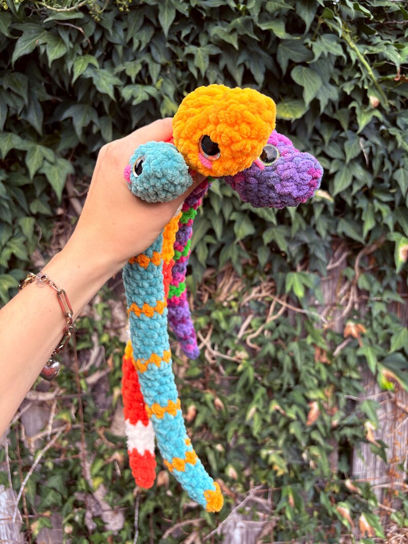 Peut inclure: Un jouet serpent en peluche &agrave; plusieurs t&ecirc;tes fabriqu&eacute; &agrave; partir de fil doux et duveteux dans des couleurs vives, notamment orange, turquoise, violet et rouge. Les trois t&ecirc;tes pr&eacute;sentent de grands yeux de s&eacute;curit&eacute; noirs brillants et des d&eacute;tails roses.