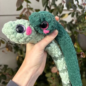 Peut inclure: Deux serpents en crochet en peluche faits de fil doux. L'un est vert menthe clair avec un nez rose et des yeux violets scintillants. L'autre est vert for&ecirc;t fonc&eacute; avec un nez rose et des yeux roses scintillants. Les deux ont des corps textur&eacute;s.