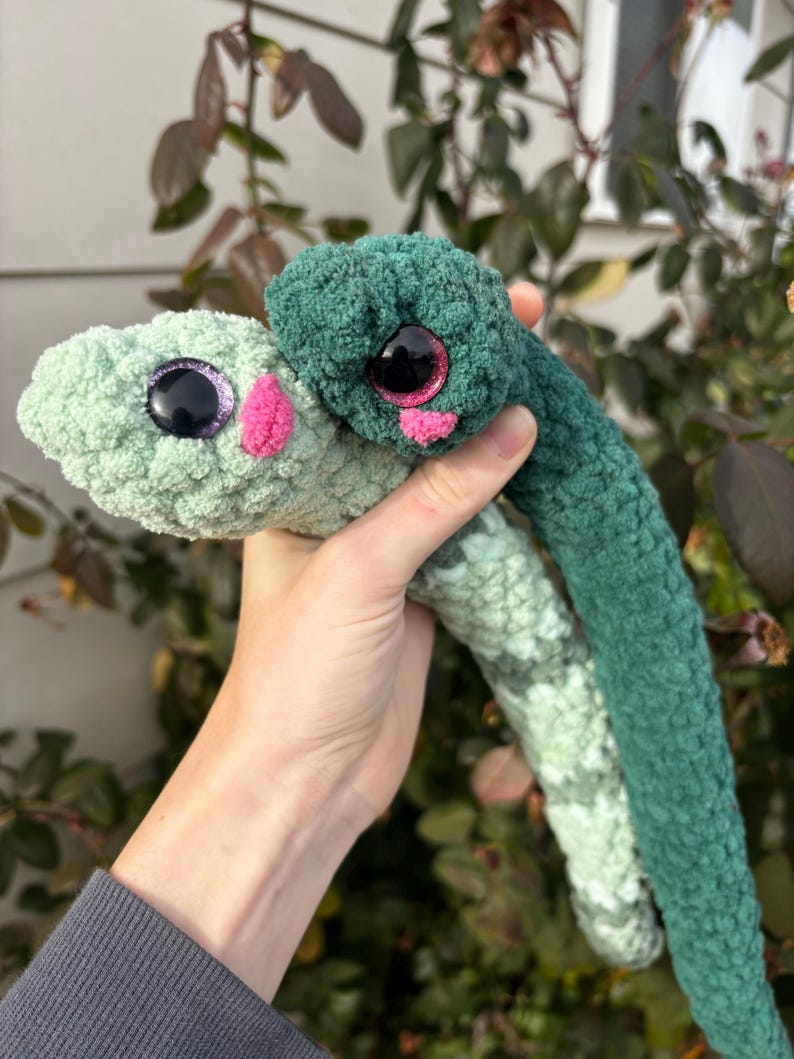 Peut inclure: Deux serpents en crochet en peluche faits de fil doux. L'un est vert menthe clair avec un nez rose et des yeux violets scintillants. L'autre est vert for&ecirc;t fonc&eacute; avec un nez rose et des yeux roses scintillants. Les deux ont des corps textur&eacute;s.