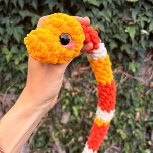 Peut inclure: Un serpent en peluche crochet&eacute; pr&eacute;sentant un motif de couleurs vives alternant orange, rouge et blanc. Le serpent poss&egrave;de une t&ecirc;te arrondie avec un grand &oelig;il en plastique noir et un petit d&eacute;tail de joue rose brod&eacute;, fait de laine textur&eacute;e.