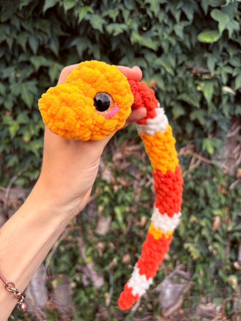 Peut inclure: Un serpent en peluche crochet&eacute; pr&eacute;sentant un motif de couleurs vives alternant orange, rouge et blanc. Le serpent poss&egrave;de une t&ecirc;te arrondie avec un grand &oelig;il en plastique noir et un petit d&eacute;tail de joue rose brod&eacute;, fait de laine textur&eacute;e.