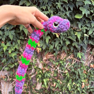 Peut inclure: Un serpent en peluche fabriqu&eacute; &agrave; partir de fil textur&eacute; doux dans un motif vibrant de violet, magenta et vert vif. Le serpent pr&eacute;sente une t&ecirc;te arrondie avec un grand &oelig;il noir brillant dot&eacute; d'un &eacute;clat iris&eacute; subtil et un long corps flexible.
