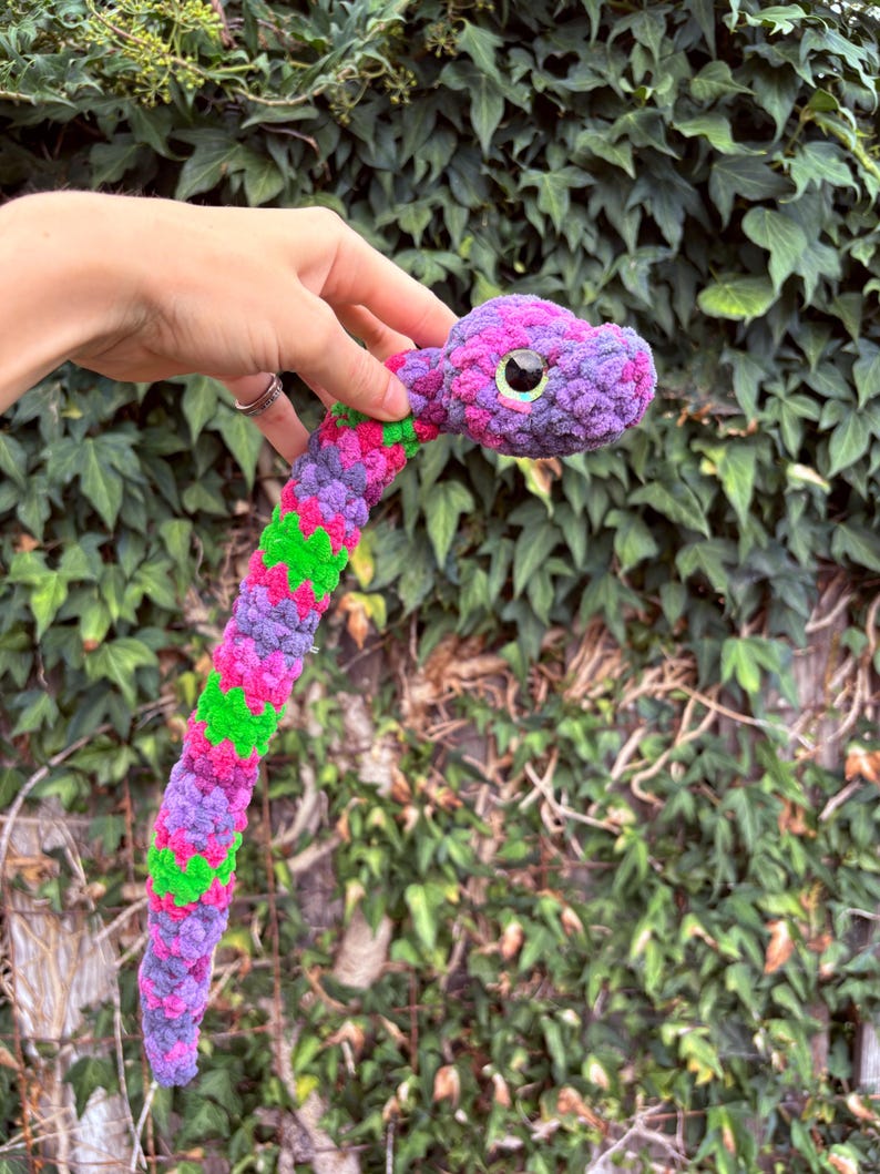 Peut inclure: Un serpent en peluche fabriqu&eacute; &agrave; partir de fil textur&eacute; doux dans un motif vibrant de violet, magenta et vert vif. Le serpent pr&eacute;sente une t&ecirc;te arrondie avec un grand &oelig;il noir brillant dot&eacute; d'un &eacute;clat iris&eacute; subtil et un long corps flexible.