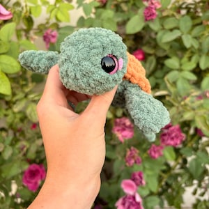 Peut inclure: Une peluche tortue crochetée en fil vert sauge doux à l'aspect texturé. La tortue présente une carapace orange clair sur le dos, de grands yeux en plastique noir avec des accents roses et quatre nageoires s'étendant de son corps.