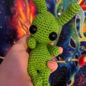Peut inclure: Un jouet en peluche extraterrestre en crochet vert avec de grands yeux noirs. Le jouet est tenu dans la main d'une personne.