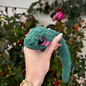 Peut inclure: Un serpent en peluche crochet&eacute; vert fonc&eacute; avec une surface en fil textur&eacute; et duveteux. Le serpent pr&eacute;sente un grand &oelig;il rond noir avec un iris &agrave; paillettes rose scintillant et un petit d&eacute;tail crochet&eacute; rose vif pr&egrave;s de la bouche.