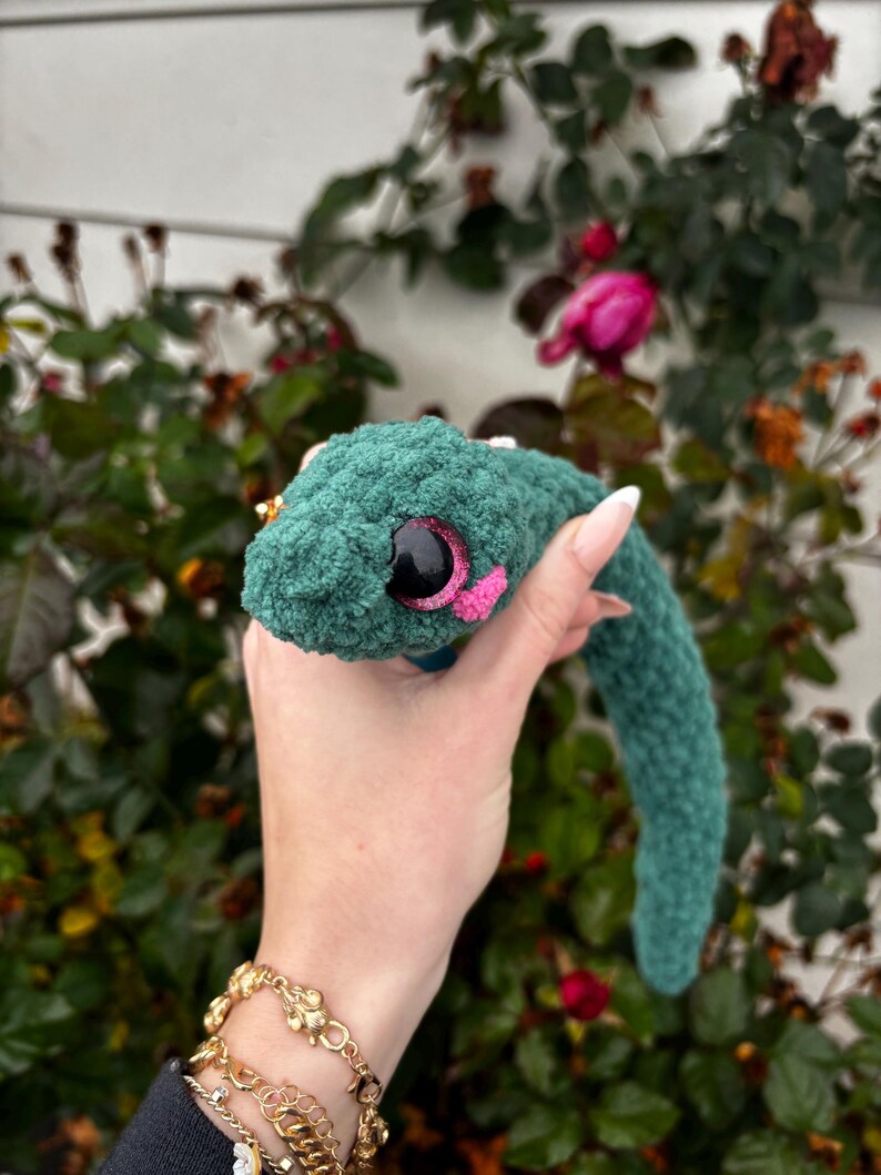 Peut inclure: Un serpent en peluche crochet&eacute; vert fonc&eacute; avec une surface en fil textur&eacute; et duveteux. Le serpent pr&eacute;sente un grand &oelig;il rond noir avec un iris &agrave; paillettes rose scintillant et un petit d&eacute;tail crochet&eacute; rose vif pr&egrave;s de la bouche.