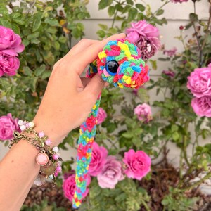 Peut inclure: Un serpent en crochet fabriqu&eacute; &agrave; partir de fil multicolore dans des tons de rose vif, jaune, vert et bleu. Le serpent pr&eacute;sente un grand &oelig;il en plastique bleu fonc&eacute; avec une pupille noire, et son corps a un aspect textur&eacute;, doux et duveteux.