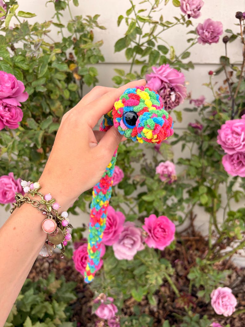 Peut inclure: Un serpent en crochet fabriqu&eacute; &agrave; partir de fil multicolore dans des tons de rose vif, jaune, vert et bleu. Le serpent pr&eacute;sente un grand &oelig;il en plastique bleu fonc&eacute; avec une pupille noire, et son corps a un aspect textur&eacute;, doux et duveteux.