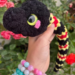 Peut inclure: Un serpent en peluche crochet&eacute; avec une t&ecirc;te noire, un grand &oelig;il jaune et noir et un petit nez en pompon rose. Le corps pr&eacute;sente un motif de bandes altern&eacute;es en laine noire, jaune et rouge avec une finition douce et textur&eacute;e.