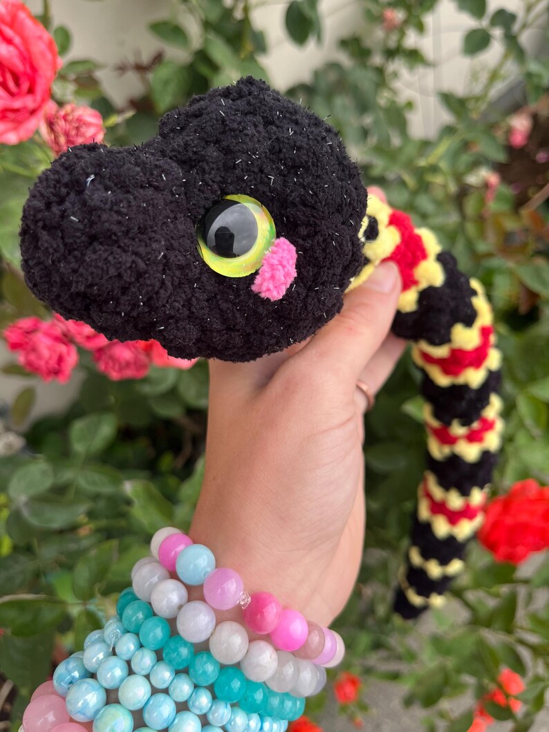 Peut inclure: Un serpent en peluche crochet&eacute; avec une t&ecirc;te noire, un grand &oelig;il jaune et noir et un petit nez en pompon rose. Le corps pr&eacute;sente un motif de bandes altern&eacute;es en laine noire, jaune et rouge avec une finition douce et textur&eacute;e.