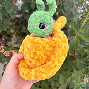 Peut inclure: Un jouet en peluche crocheté à la main présentant une tête verte semblable à une chenille avec deux antennes et de grands yeux noirs, niché dans une base arrondie texturée d'un orange vif. Le fil doux et duveteux crée un aspect pelucheux.