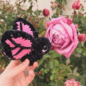 Peut inclure: Un papillon en peluche fabriqué avec du fil doux noir et rose vif. Le papillon présente un corps arrondi, de grandes ailes avec un motif noir et rose distinct, et un seul œil noir brillant visible. La texture est duveteuse et veloutée.