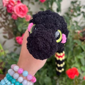 Peut inclure: Un serpent en peluche crochet&eacute; noir avec une surface textur&eacute;e, de grands yeux jaunes et noirs, et des taches roses sur les joues. Le corps pr&eacute;sente un motif r&eacute;p&eacute;titif de segments jaunes et rouges. La laine est duveteuse avec des taches blanches.