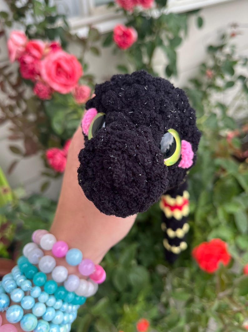 Peut inclure: Un serpent en peluche crochet&eacute; noir avec une surface textur&eacute;e, de grands yeux jaunes et noirs, et des taches roses sur les joues. Le corps pr&eacute;sente un motif r&eacute;p&eacute;titif de segments jaunes et rouges. La laine est duveteuse avec des taches blanches.