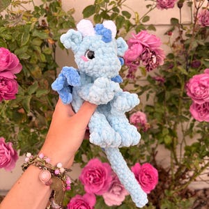 Peut inclure: Une peluche dragon au crochet bleu clair faite de fil doux et texturé. La créature présente des cornes blanches et bleues, de petites ailes, une longue queue et de grands yeux noirs avec des touches roses sur les joues, montrant une fabrication artisanale détaillée.