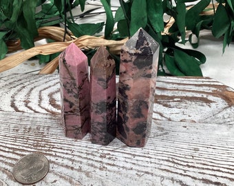 Puede incluir: Tres obeliscos de cristal de rodonita pulidos. Los cristales son una mezcla de rosa y negro, con un acabado pulido. Los obeliscos están de pie sobre una superficie de madera blanca. Los cristales varían en tamaño y forma.