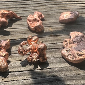 May include: Six irregularly shaped copper nuggets with a textured surface are displayed on a weathered wooden surface. The nuggets vary in size and shape, with a reddish-brown color. The lighting creates shadows, highlighting the uneven surfaces.