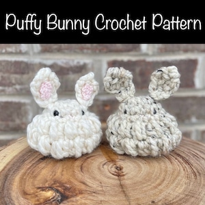 May include: Two crocheted bunny plushies, one white and one gray, with pink ears. The bunnies are sitting on a wooden surface.  Puffy Bunny Crochet Pattern.