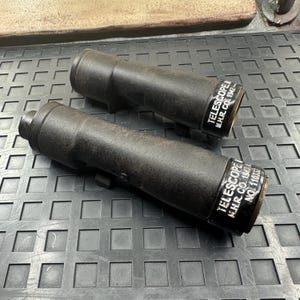 May include: Two vintage black telescopes with brass-colored ends are displayed on a dark surface. Each telescope has the text "TELESCOPE I M.H.R. CO. 1942" printed on the end. The telescopes are cylindrical in shape.