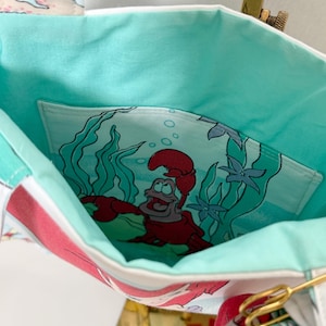 Puede incluir: Un bolso de tela con forro interior de color turquesa claro y un bolsillo interior profundo. El bolsillo muestra un estampado gr&aacute;fico de un personaje de cangrejo rojo rodeado de algas verdes y agua azul. El exterior muestra un patr&oacute;n de coral.
