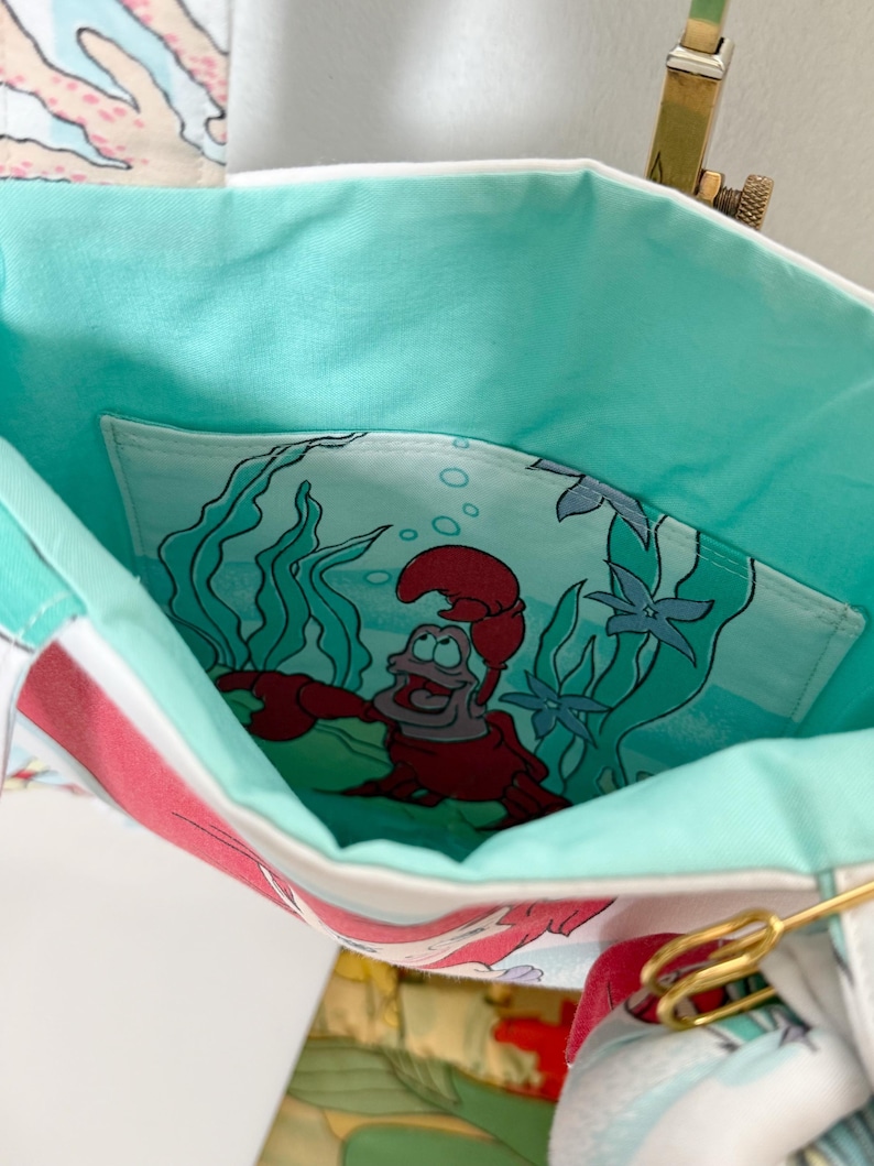 Puede incluir: Un bolso de tela con forro interior de color turquesa claro y un bolsillo interior profundo. El bolsillo muestra un estampado gr&aacute;fico de un personaje de cangrejo rojo rodeado de algas verdes y agua azul. El exterior muestra un patr&oacute;n de coral.