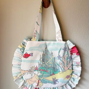 Puede incluir: Un bolso de tela blanco con una colorida escena submarina que muestra un gran castillo, un rey barbudo con un tridente y varios animales marinos. El bolso tiene forma redonda con un borde con volantes y dos asas de la misma tela.