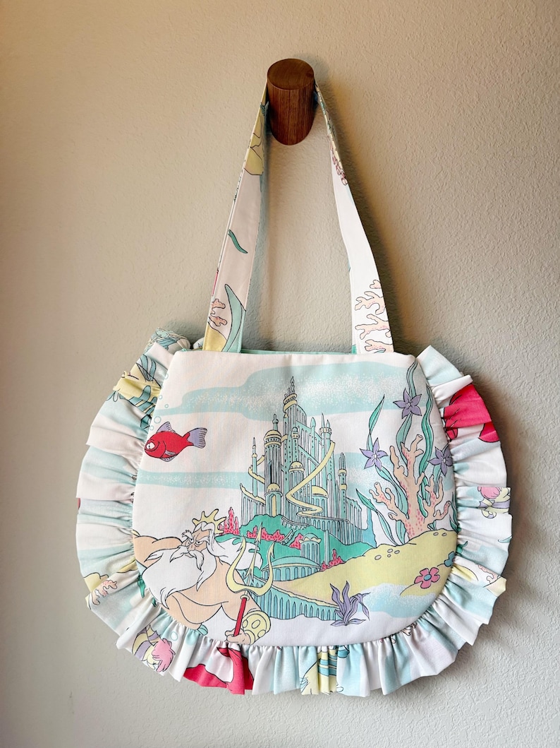 Puede incluir: Un bolso de tela blanco con una colorida escena submarina que muestra un gran castillo, un rey barbudo con un tridente y varios animales marinos. El bolso tiene forma redonda con un borde con volantes y dos asas de la misma tela.