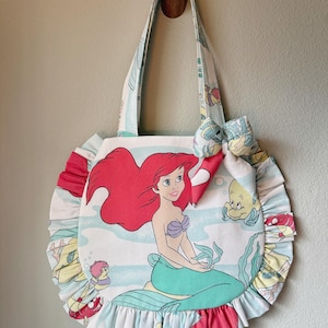 Puede incluir: Un bolso de tela redondo con un estampado de una sirena de pelo rojo con cola verde y un pez amarillo. El bolso tiene un fondo azul claro con elementos submarinos, un borde con volantes y un lazo de tela decorativo en la correa.