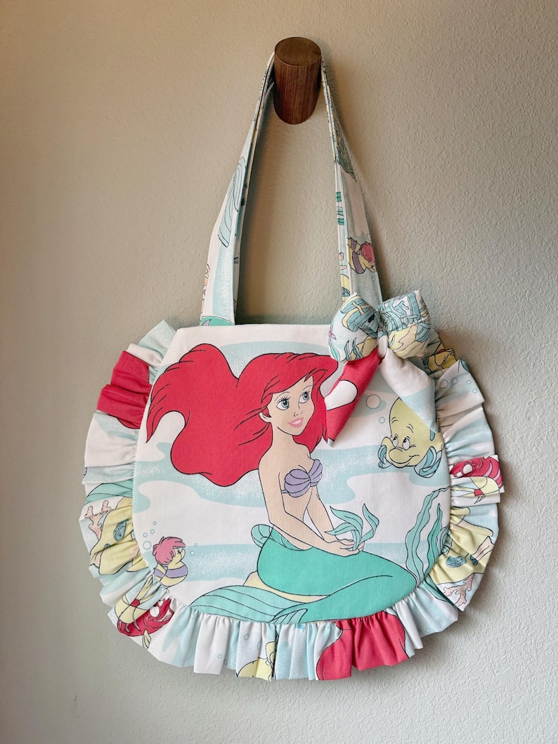 Puede incluir: Un bolso de tela redondo con un estampado de una sirena de pelo rojo con cola verde y un pez amarillo. El bolso tiene un fondo azul claro con elementos submarinos, un borde con volantes y un lazo de tela decorativo en la correa.