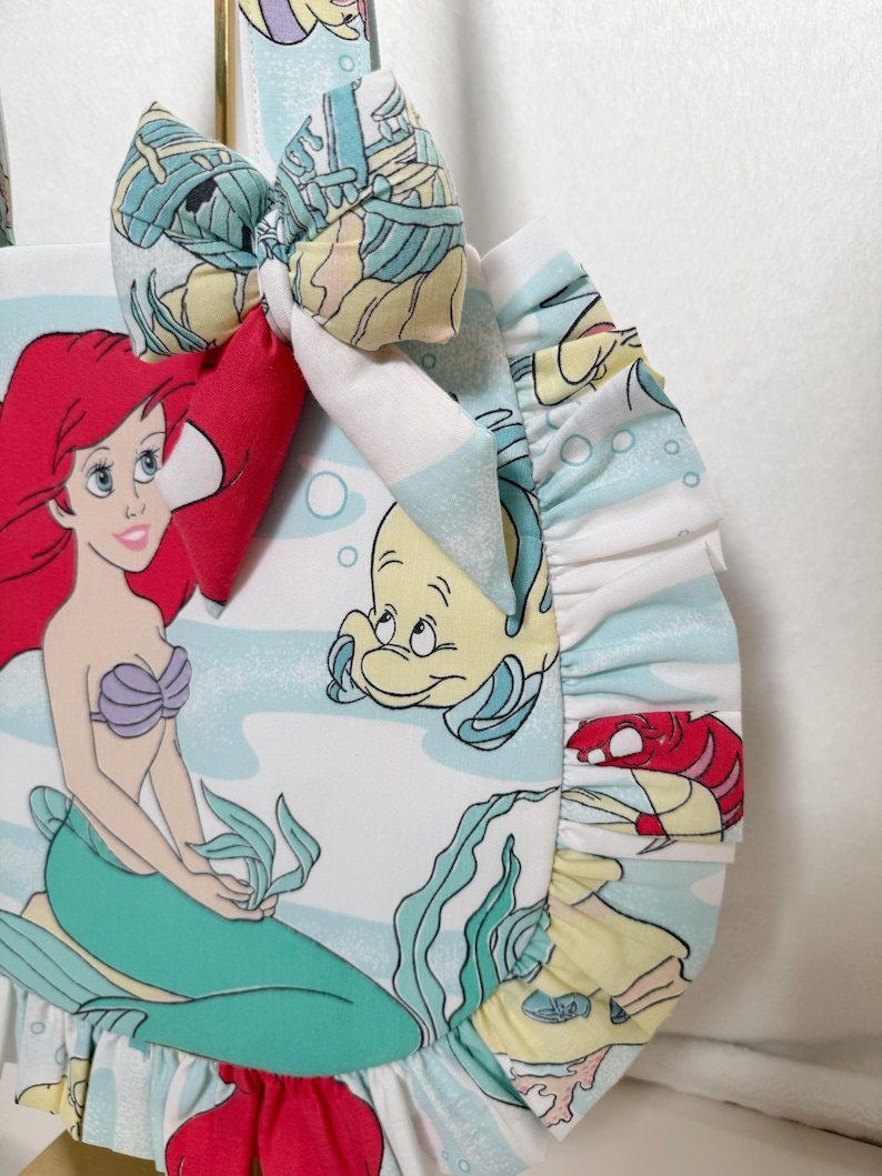Puede incluir: Un bolso de tela redondo con un estampado de una sirena de pelo rojo, un pez amarillo y un cangrejo rojo sobre un fondo azul claro con burbujas. El bolso tiene un borde con volantes, un lazo de tela a juego y un asa de tela estampada.