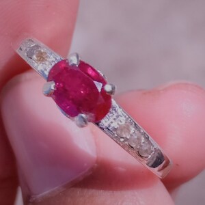 Peut inclure: Une bague en argent ornée d'un rubis ovale vif en son centre. La monture est ornée de petits diamants transparents. La bague est présentée sur un fond neutre, mettant en valeur son design élégant.