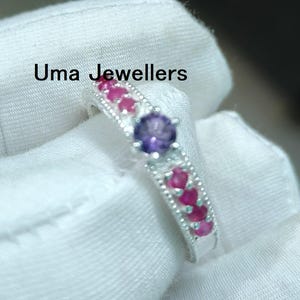 May include: A silver ring featuring a central purple gemstone, flanked by smaller pink gemstones. The ring is held in a gloved hand, with the text "Uma Jewellers" visible.