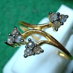 Op de afbeelding: Een goudkleurige ring met een bloemmotief. De ring heeft clusters van kleine, ronde, lichtgekleurde stenen. De band buigt om de vinger, met de bloemelementen bovenop. De ring wordt gepresenteerd op een witte standaard tegen een groene achtergrond.