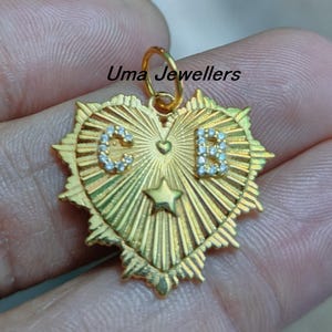 May include: A gold-colored pendant in the shape of a heart with a star and the letters "C" and "B" encrusted with small white stones. The pendant has a sunburst design and the text "Uma Jewellers".