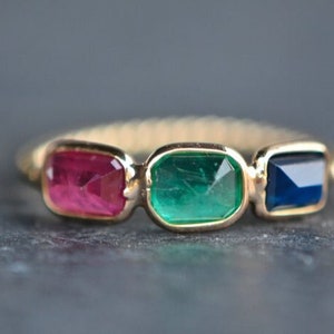 May include: A gold ring with three rectangular gemstones: a red ruby, a green emerald, and a blue sapphire.
