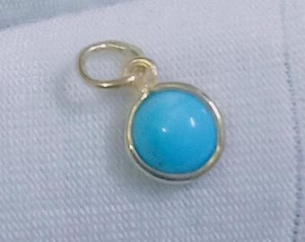 May include: A small, round pendant with a bright turquoise stone set in a gold-colored metal. The pendant has a simple design with a loop at the top for a chain. The background is a white, textured fabric.
