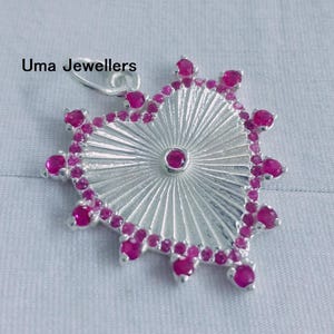 Könnte beinhalten: Ein silberner Anhänger mit Sonnenstrahlen-Design, verziert mit zahlreichen kleinen, leuchtend rosa Edelsteinen. Der Anhänger hat einen zentralen Edelstein und ist herzförmig. Der Text "Uma Jewellers" ist sichtbar.