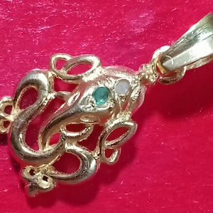 May include: A gold-colored pendant in the shape of the Hindu deity Ganesha, with an emerald and a small white gemstone. The pendant has intricate details and a loop for a chain. The background is red.