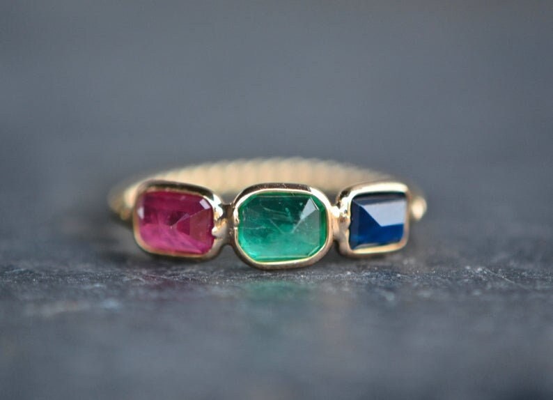 May include: A gold ring with three rectangular gemstones: a red ruby, a green emerald, and a blue sapphire.