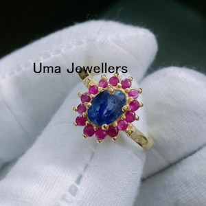 May include: A gold ring featuring a central oval blue sapphire surrounded by a halo of small round pink rubies. The text Uma Jewellers is visible above the ring. The piece is set in a polished yellow gold band with a classic prong setting design.