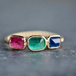 May include: A gold ring with three rectangular gemstones: a red ruby, a green emerald, and a blue sapphire.