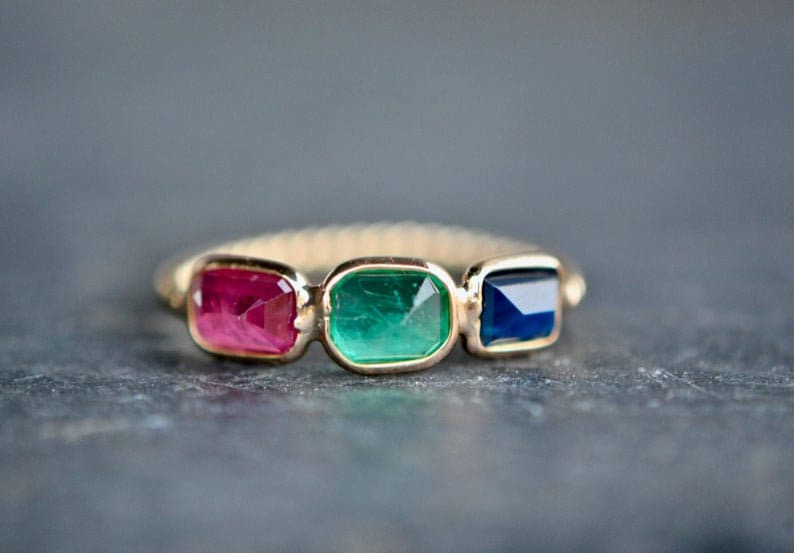 May include: A gold ring with three rectangular gemstones: a red ruby, a green emerald, and a blue sapphire.
