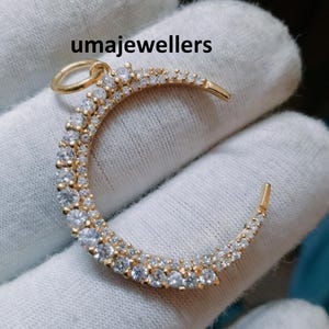 May include: A gold crescent moon pendant with a double row of clear cubic zirconia stones. The pendant is on a white background.