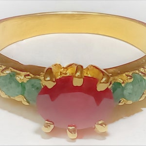 May include: A gold ring featuring a large red gemstone surrounded by smaller green gemstones.