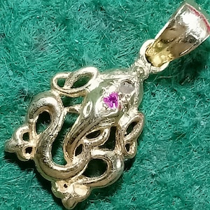 May include: A gold-colored pendant featuring an ornate design with a central figure and swirling details. A small, round, pink gemstone is set into the pendant. The pendant is displayed against a green background.