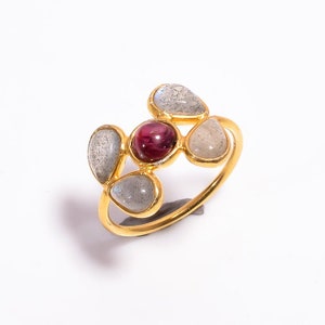 Può includere: Un anello dorato con una gemma centrale rotonda di colore rosso scuro, circondata da quattro gemme grigie a forma di goccia. Il design dell'anello è ispirato ai fiori, con le gemme disposte a grappolo.