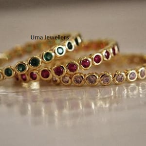 May include: Three gold bangles with a row of small, colorful gemstones. The bangles are stacked on top of each other. The top bangle has green gemstones, the middle bangle has red gemstones, and the bottom bangle has clear gemstones.